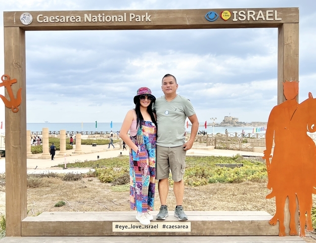       Two people posing under a wooden frame with a distant sea scene.
  