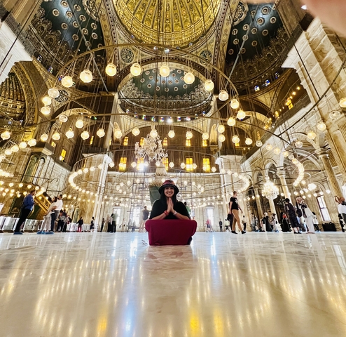 Person sitting in an illuminated mosque with hanging lights.