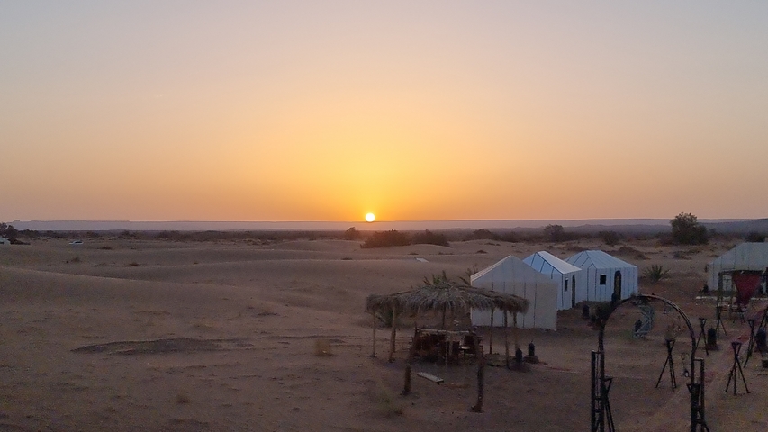 Desert landscape at sunset with tents and a palm-covered shelter.
