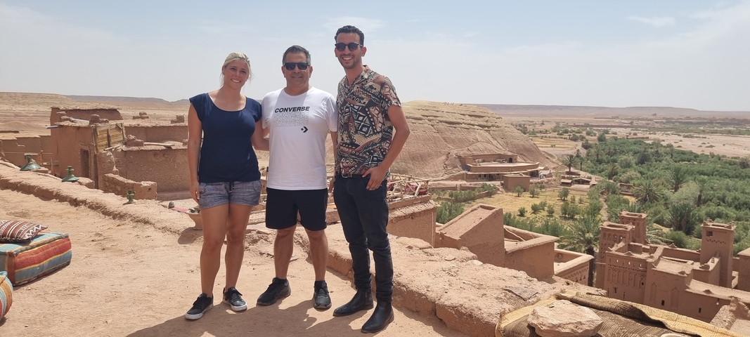 Three people posing in front of desert landscape with mud-brick buildings.
