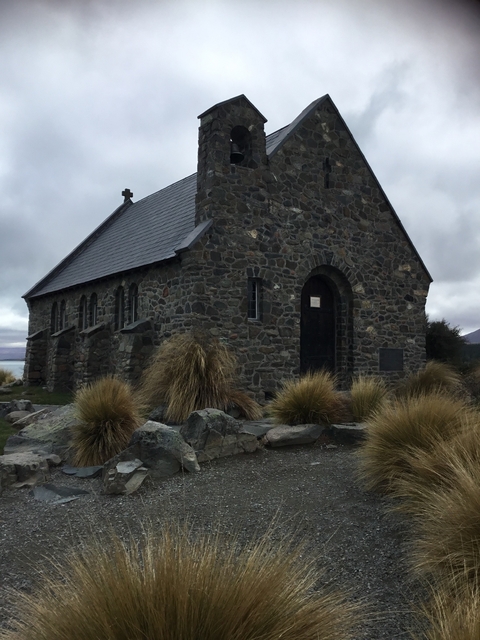       A stone church building surrounded by dry plants under a cloudy sky.
  