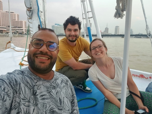 Three people smiling on a boat with a city skyline in the background.