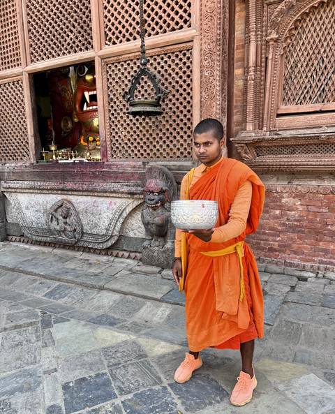       A monk in orange robes holding a bowl beside traditional architecture.
  