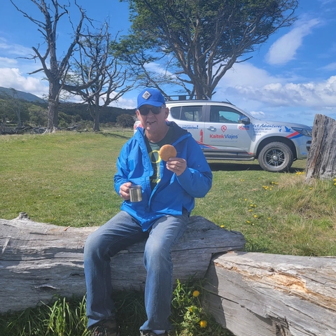       Man sitting on a log with a snack and a vehicle in the background.
  