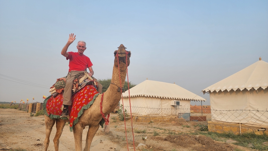 Man waving while riding a camel near tents.