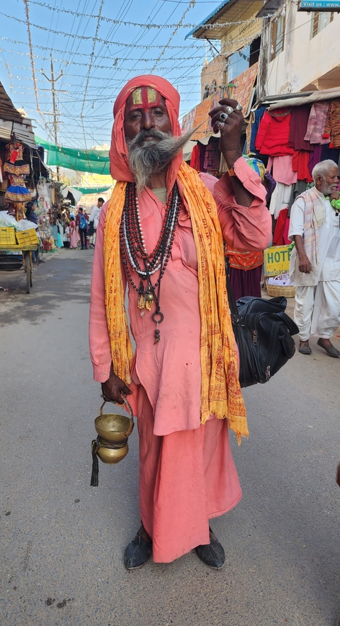       A man in traditional attire with beads and a scarf, standing in a bustling street.
  