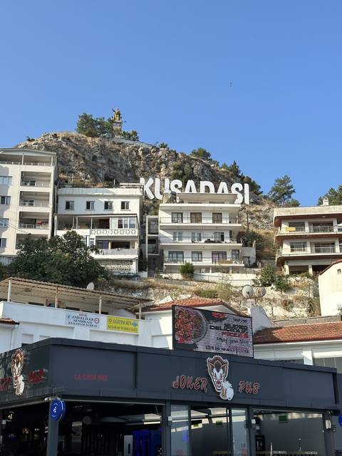       Kusadasi sign on a hill with shops beneath.
  