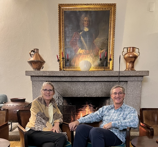 Two people sitting in front of a fireplace with a painting above.