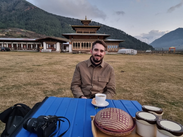       Man sitting outdoors with tea in front of a traditional Bhutanese structure.
  