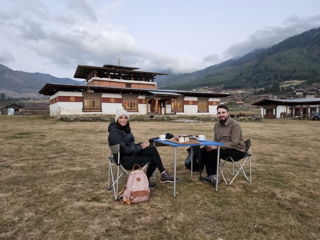       Couple sitting outdoors at a table with a traditional building in the background.
  