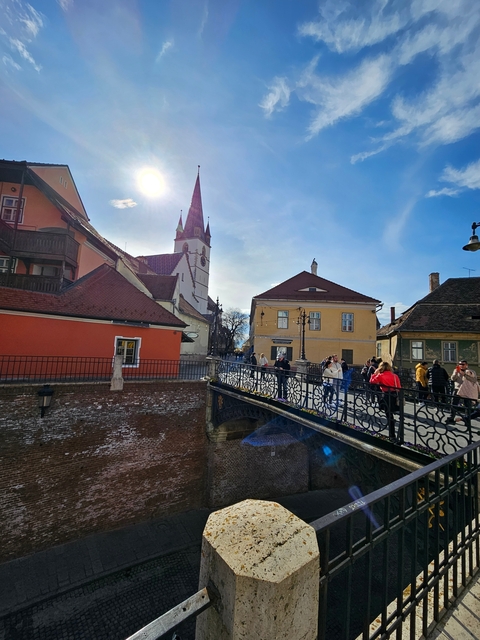 Street scene of a European town with people walking on a bridge, colorful buildings, and a church tower.