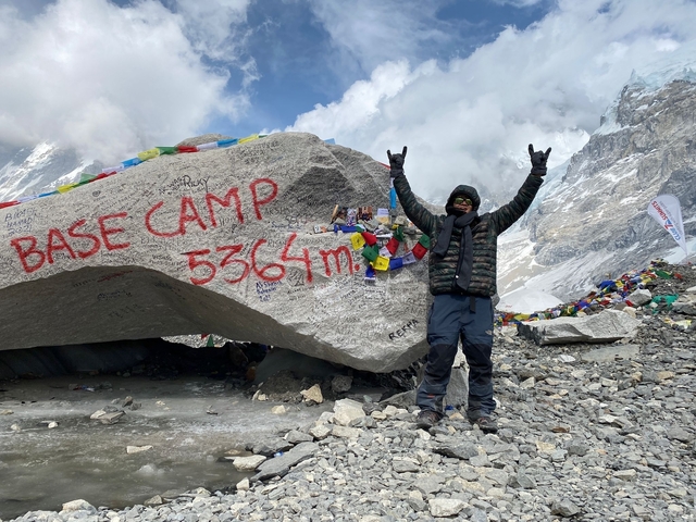 Person posing next to a rock with 'Base Camp 5364m' written on it surrounded by mountains.