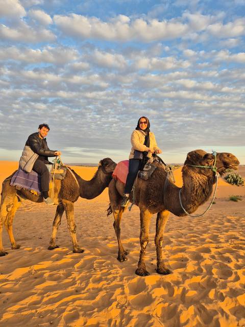 Two people riding camels on sand dunes during sunset.