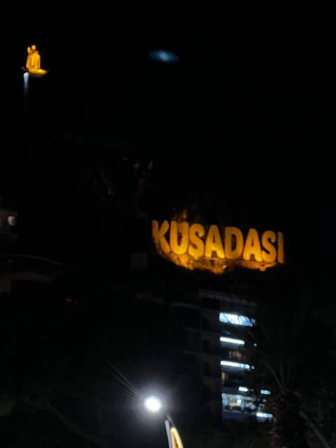 A night scene with an illuminated sign that says 'Kusadasi'.