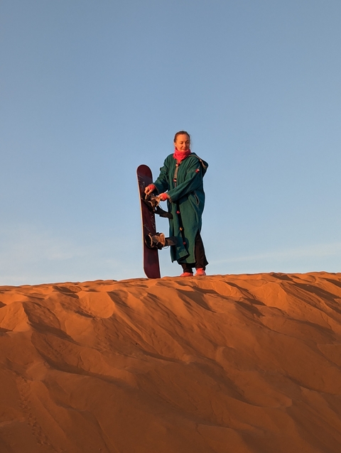       A person dressed in traditional clothing holding a sandboard on a sand dune.
  