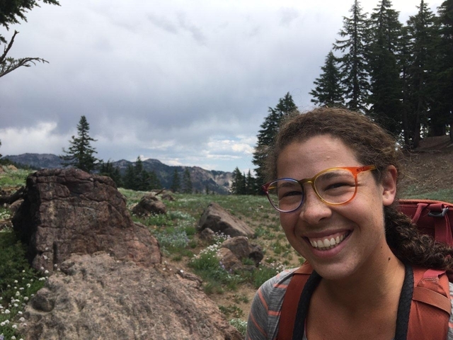       Smiling person wearing glasses with a mountainous landscape background.
  