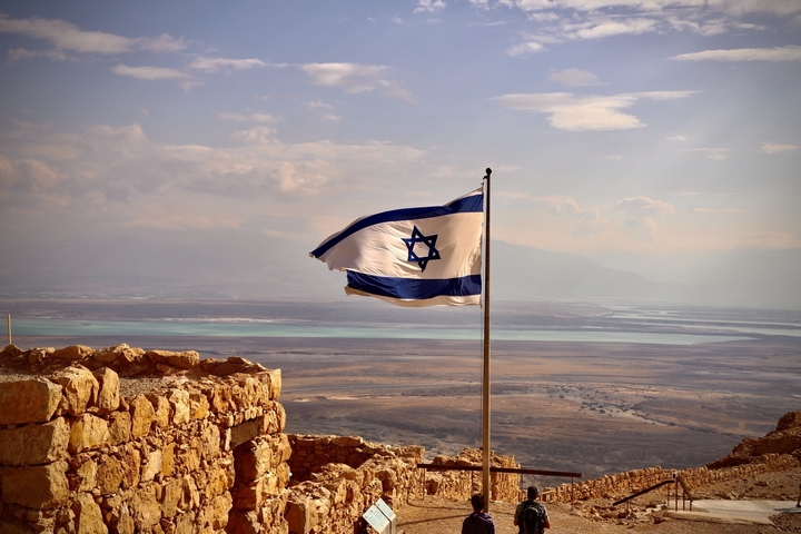       Israeli flag viewed over a landscape with distant mountains.
  