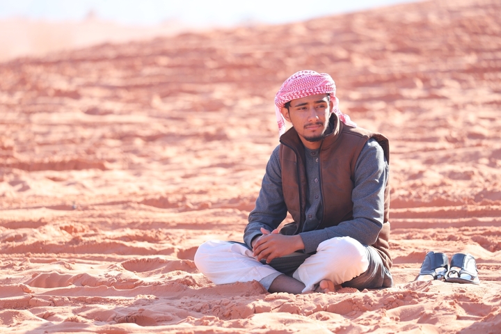       Man sitting on desert sand wearing traditional clothing.
  