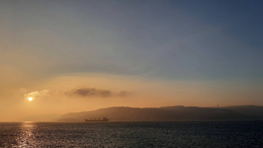       Sunset over a body of water with a cargo ship and distant hills.
  