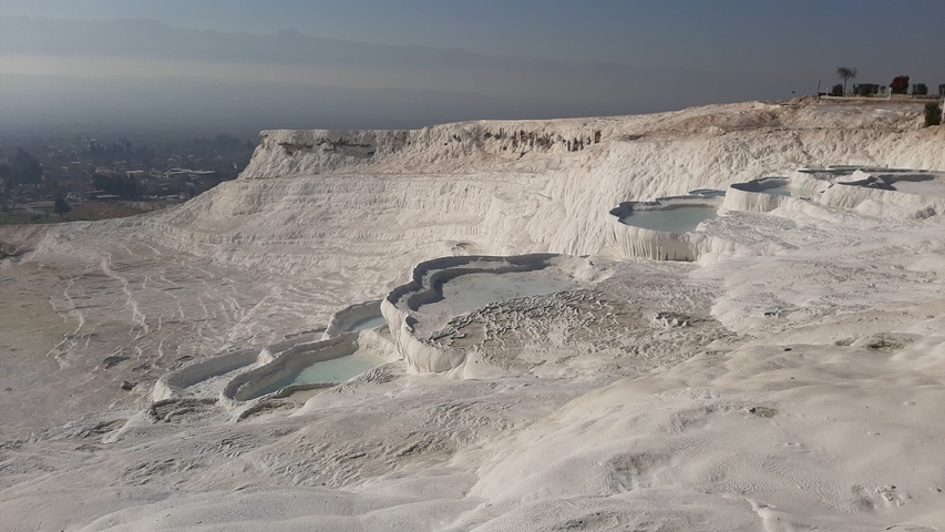       Pamukkale's travertine terraces filled with water under a hazy sky.
  