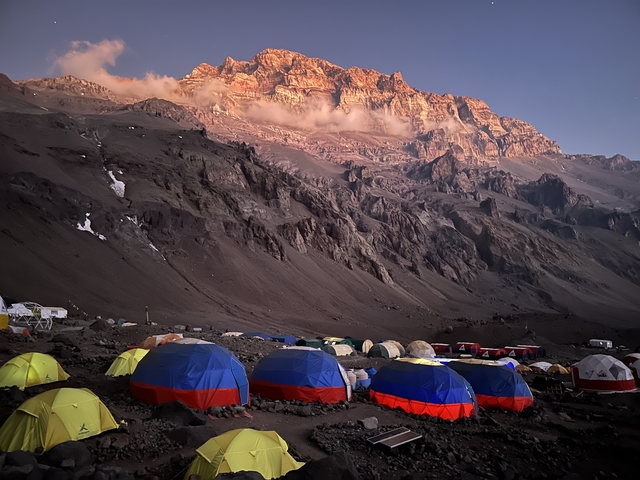       Mountain campsite with colorful tents and a rocky peak at sunrise.
  