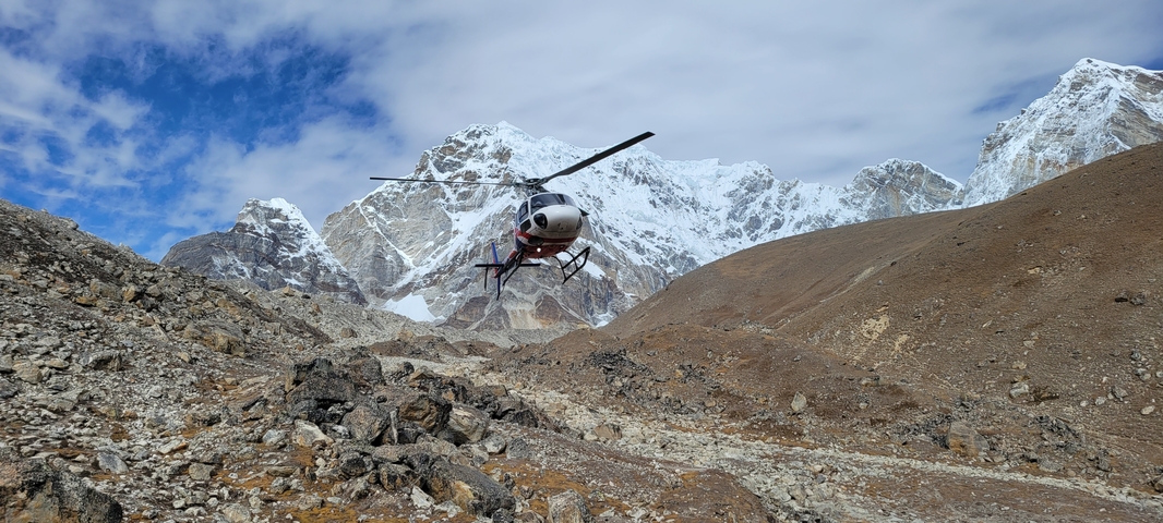       Helicopter flying over rocky terrain with snowy mountains.
  