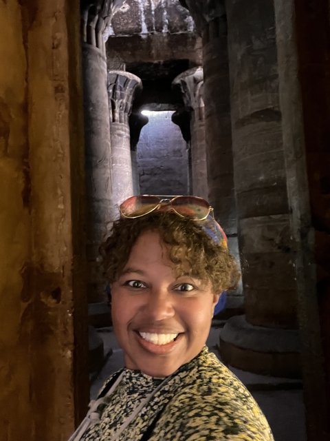       Person smiling indoors beside ancient ruins.
  