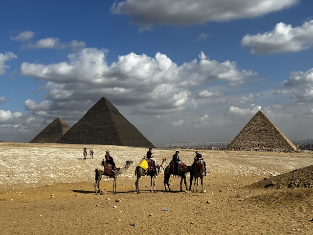       Camels and riders near the pyramids with cloudy skies.
  