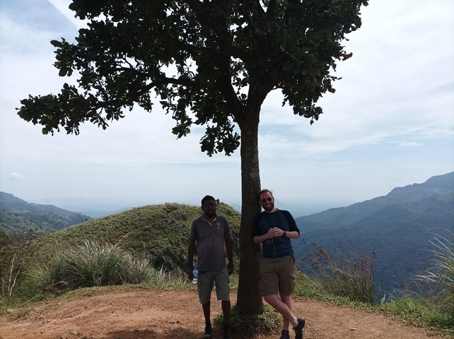 Two people standing under a tree with scenic hills in the background.