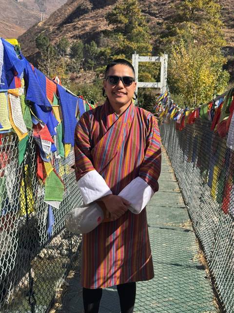 A person in traditional Bhutanese attire standing on a bridge with prayer flags.