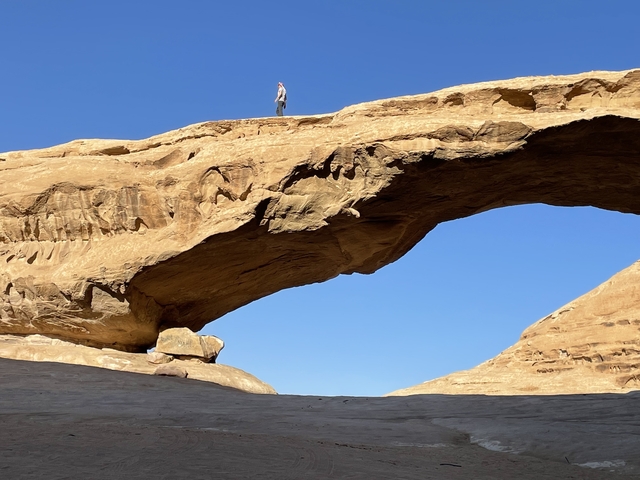Person standing on top of a large natural rock arch in a desert landscape.