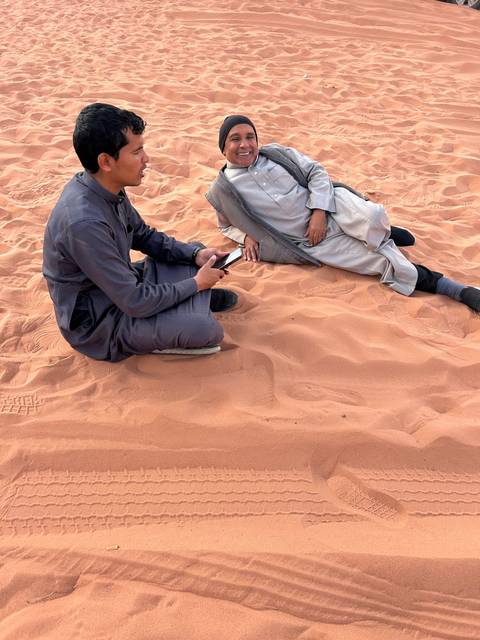Two boys sitting on red sand with impressions and shadows.