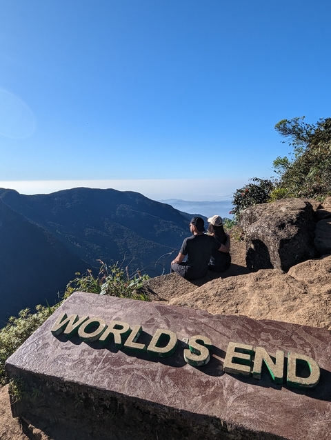       Two people sitting on a cliff with expansive views of mountains.
  
