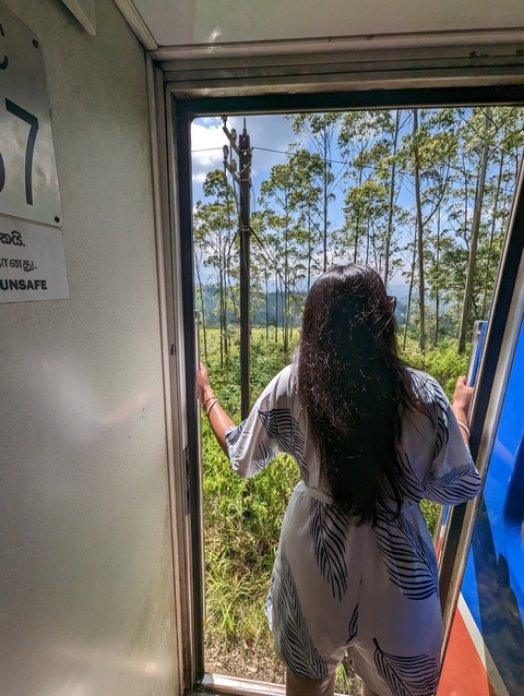      Person leaning out of a train door with lush greenery outside.
  