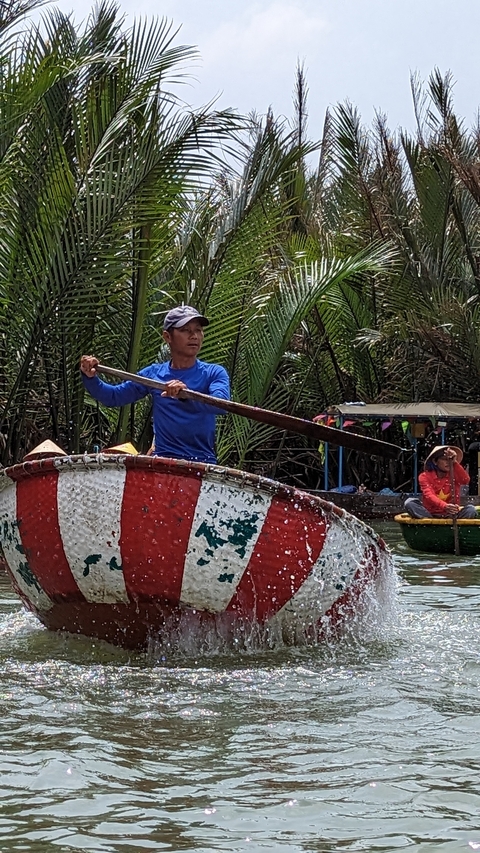       Man rowing a basket boat on a river with trees in the background.
  