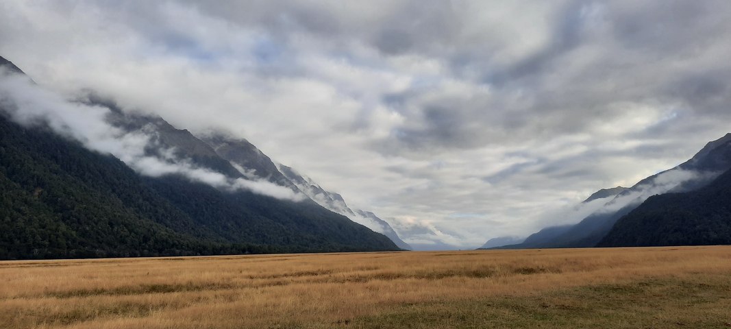 Vast valley with clouds hanging low over mountains and grassy fields.