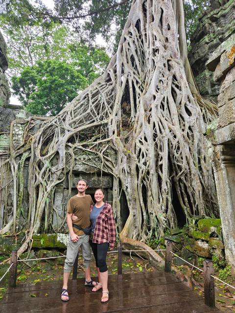 Two people in front of large tree roots growing over temple ruins.