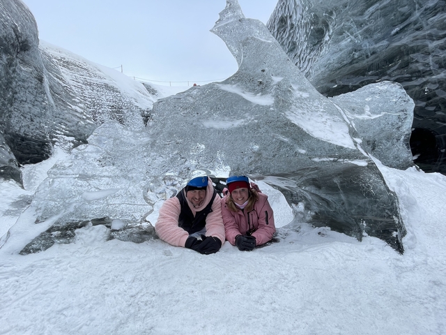       Two people lying under a natural ice arch in the snow.
  