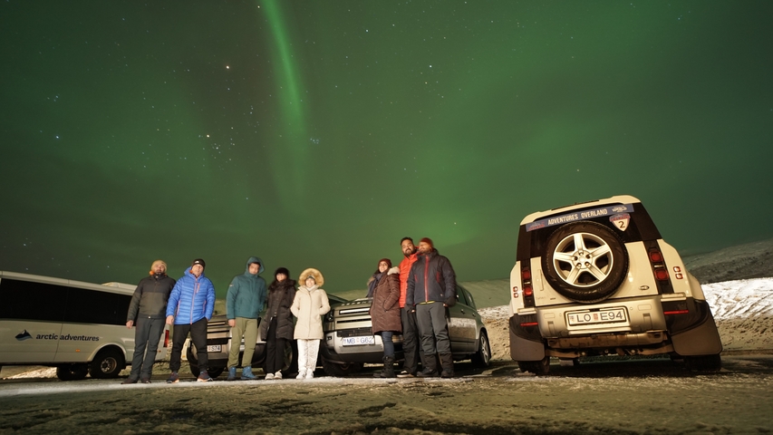 People standing under the northern lights with cars nearby.