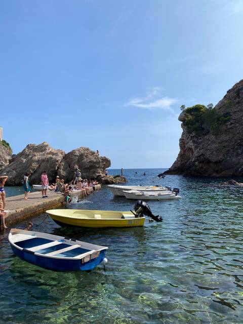       People relaxing on boats and rocks in a coastal area with clear water and rocky formations.
  