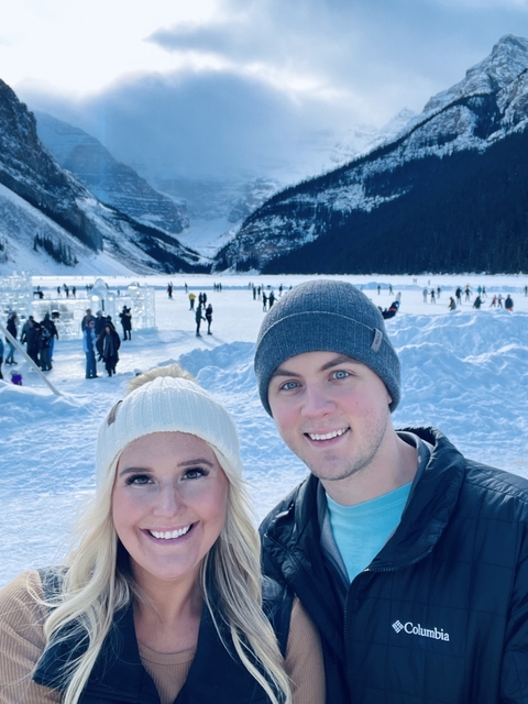       A couple smiles in a snowy setting with distant ice sculptures.
  