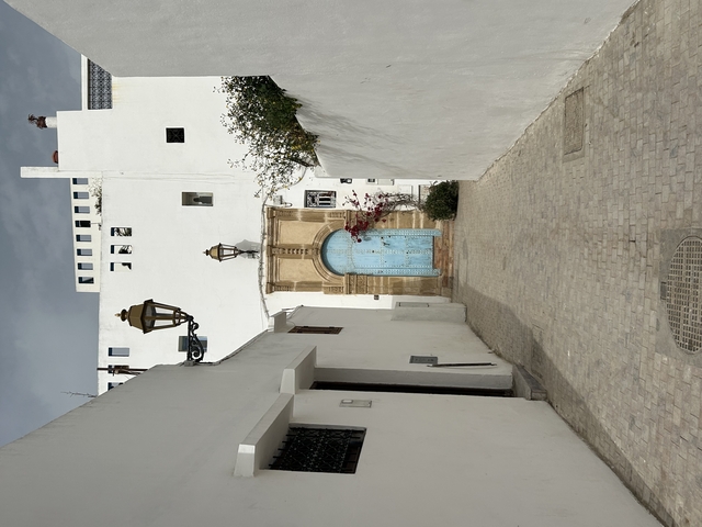       White buildings with a large blue fountain in the center of a narrow street.
  