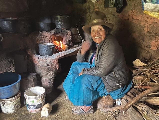 Elderly woman sitting by a rustic hearth with pots.