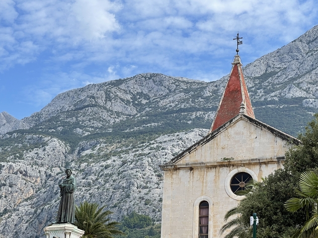       Church with a red rooftop and statue set against a mountain backdrop.
  