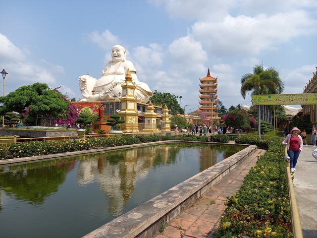 Large white Buddha statue in a temple garden with a pond and people around.