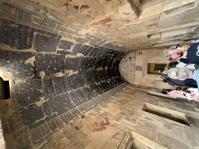       Interior of an ancient structure with ornate ceiling and people walking.
  