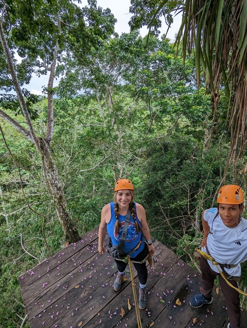       Two people with helmets, hanging at height among trees on an adventure course.
  