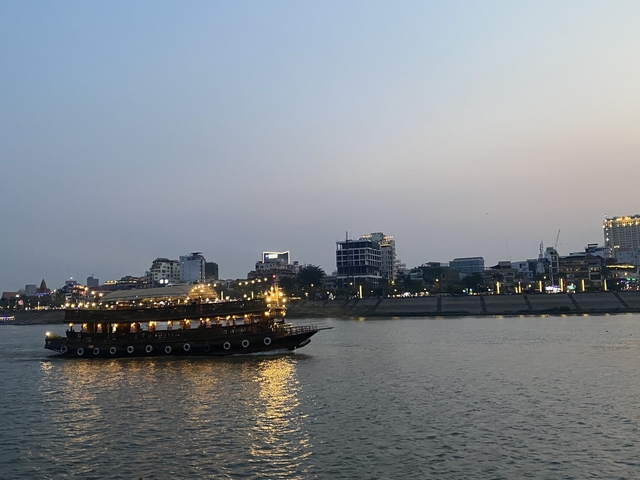 A lit tourist boat cruising along a river with cityscape in the background during dusk.