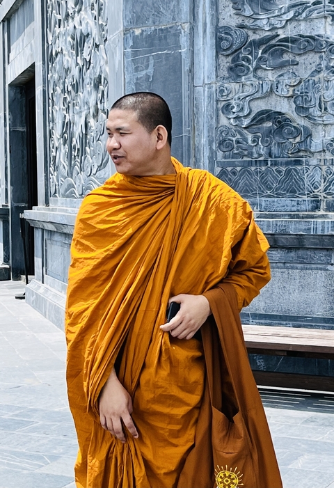 A person in an orange robe standing against a textured stone wall.