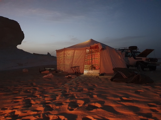 A tent lit up at night in a desert area with a vehicle nearby.